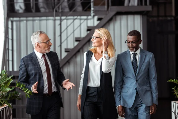 Handsome businessman in glasses gesturing while looking at attractive woman smiling near african american partner — Stock Photo