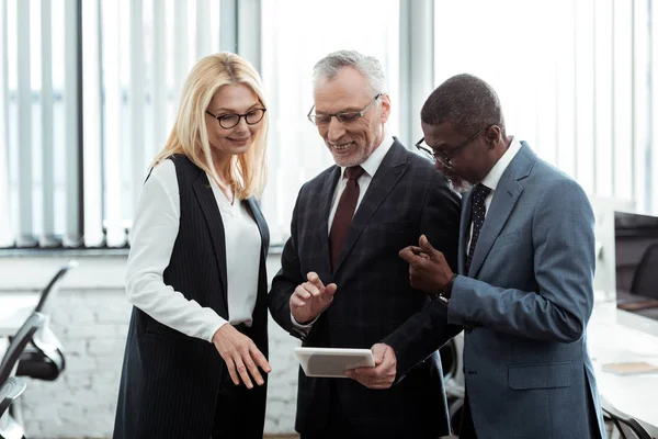 Happy blonde businesswoman looking at digital tablet near multicultural businessmen — Stock Photo
