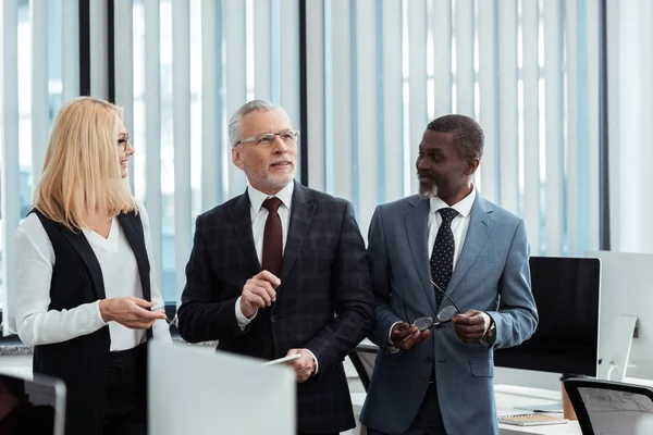 Glückliche blonde Frau schaut Geschäftsmann mit Brille in der Nähe eines afrikanisch-amerikanischen Mannes an — Stockfoto