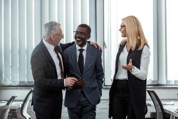 Feliz hombre de negocios abrazando guapo africano americano compañero de trabajo cerca de mujer de negocios - foto de stock