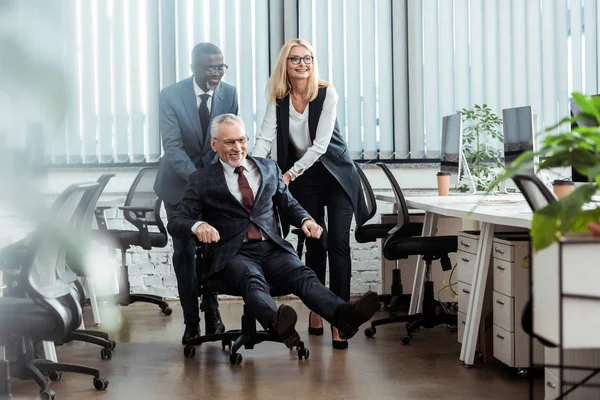 Selektiver Fokus des glücklichen Geschäftsmannes in Brille auf Stuhl neben multikulturellen Mitarbeitern — Stockfoto