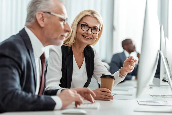 Enfoque selectivo de rubia sonriente mujer de negocios en gafas apuntando con el dedo mientras mira a su compañero de trabajo - foto de stock