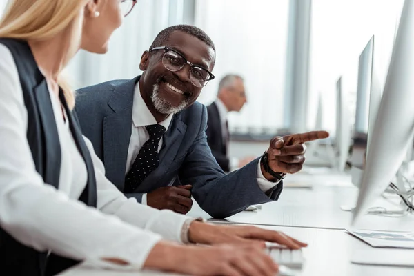 Selektiver Fokus des fröhlichen afrikanisch-amerikanischen Geschäftsmannes, der mit dem Finger auf den Computermonitor in der Nähe eines blonden Kollegen zeigt — Stockfoto