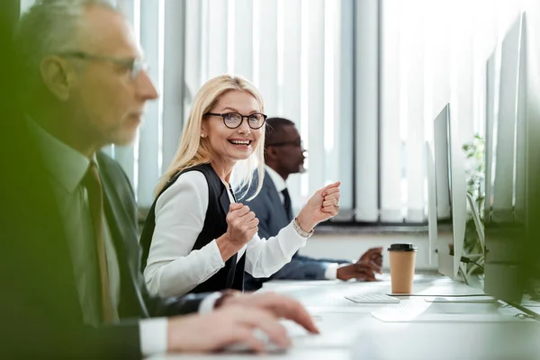 Foco seletivo de mulher de negócios loira feliz sorrindo e gesticulando perto de homens no escritório — Fotografia de Stock