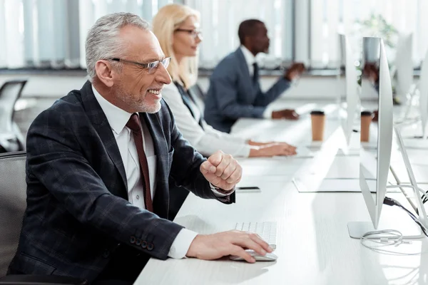 Selective focus of happy businessman smiling and gesturing near multicultural coworkers in office — Stock Photo