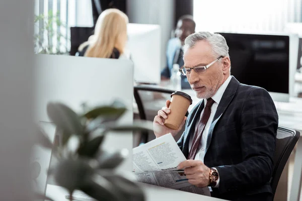 Selective focus of handsome businessman reading newspaper while holding disposable cup near multicultural coworkers — Stock Photo