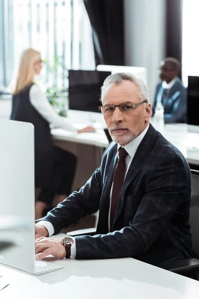 Selektiver Fokus des Geschäftsmannes in Brille, der in die Kamera in der Nähe multikultureller Mitarbeiter blickt — Stockfoto