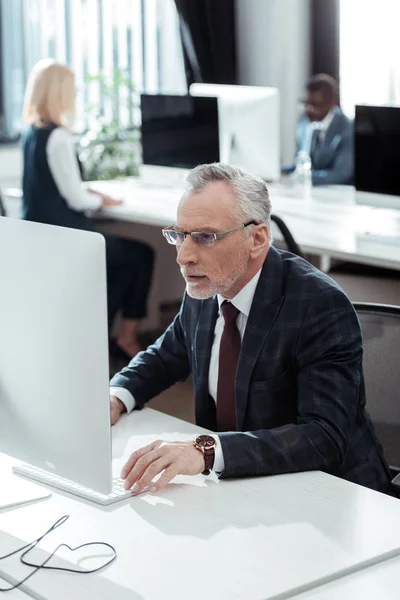 Selective focus of handsome mature businessman in glasses looking at computer monitor near multicultural colleagues in office — Stock Photo