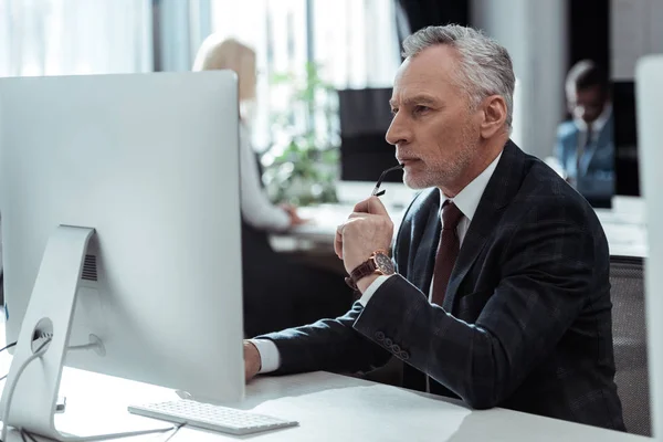 Enfoque selectivo de empresario maduro pensativo sosteniendo gafas y mirando el monitor de la computadora cerca de colegas multiculturales en la oficina - foto de stock