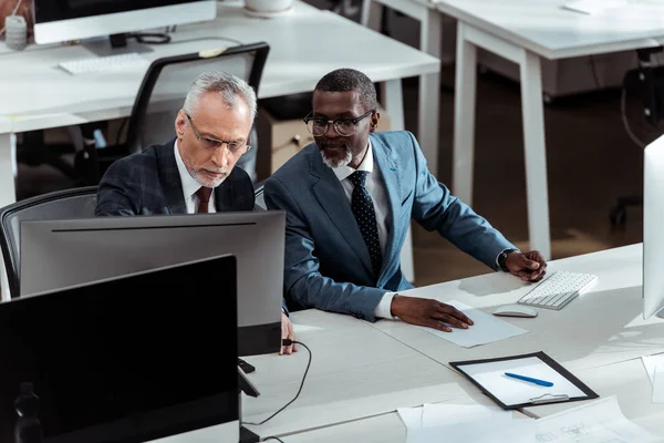 Gutaussehender afrikanisch-amerikanischer Geschäftsmann schaut auf Computermonitor in der Nähe seines Büromitarbeiters — Stockfoto