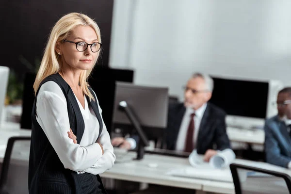 Selective focus of attractive businesswoman in glasses standing with crossed arms near multicultural men — Stock Photo