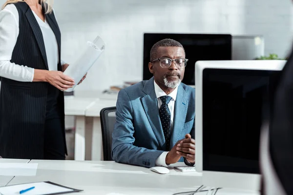 Vista recortada de mujer de negocios sosteniendo planos cerca de hombre de negocios afroamericano en gafas - foto de stock