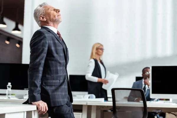 Selective focus of handsome businessman standing with closed eyes near multicultural coworkers — Stock Photo