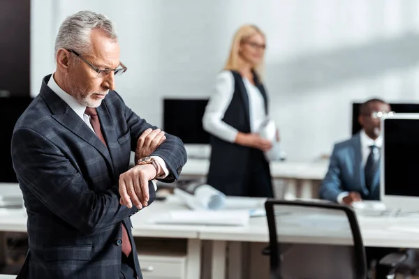 Selective focus of handsome businessman looking at watch near multicultural coworkers — Stock Photo