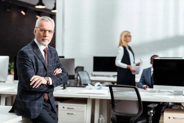Selective focus of businessman in glasses looking at camera while standing with crossed arms in office — Stock Photo