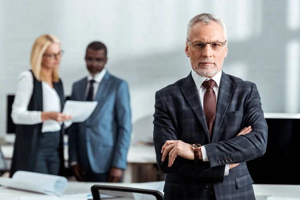 Selective focus of confident businessman in glasses and looking at camera while standing with crossed arms near colleagues — Stock Photo