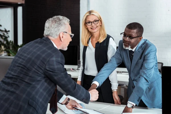 Enfoque selectivo de sonriente mujer de negocios rubia cerca de hombres de negocios multiculturales - foto de stock