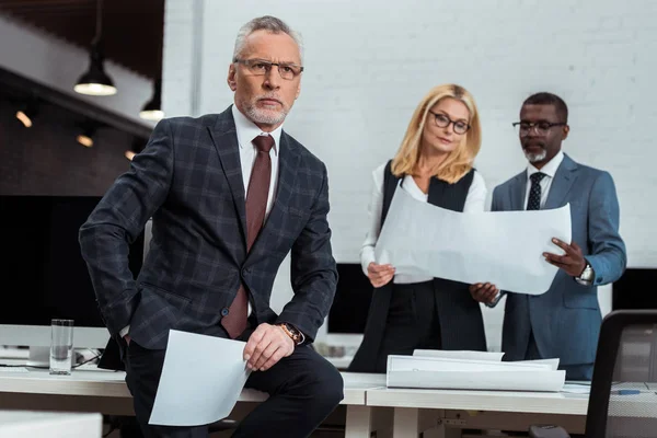 Selective focus of thoughtful businessman holding blank paper near multicultural partners standing with blueprints — Stock Photo