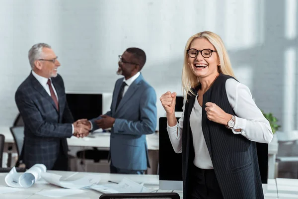 Selective focus of happy businesswoman celebrating triumph near multicultural businessmen shaking hands in office — Stock Photo