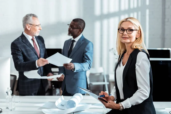 Enfoque selectivo de la mujer de negocios en gafas sujetando portapapeles cerca de hombres de negocios multiculturales - foto de stock