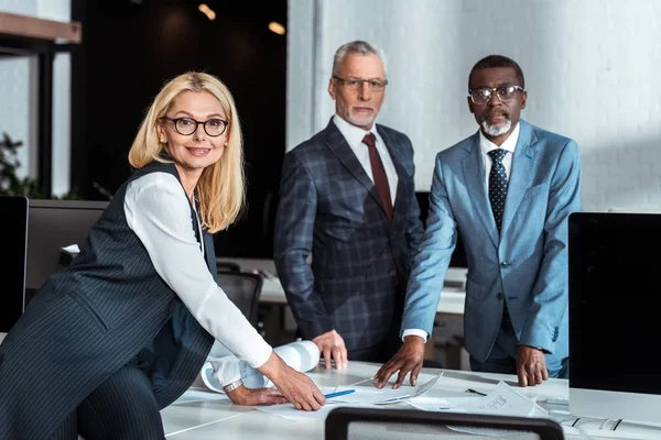 Enfoque selectivo de mujer de negocios rubia en gafas de pie cerca de la mesa y hombres de negocios multiculturales - foto de stock