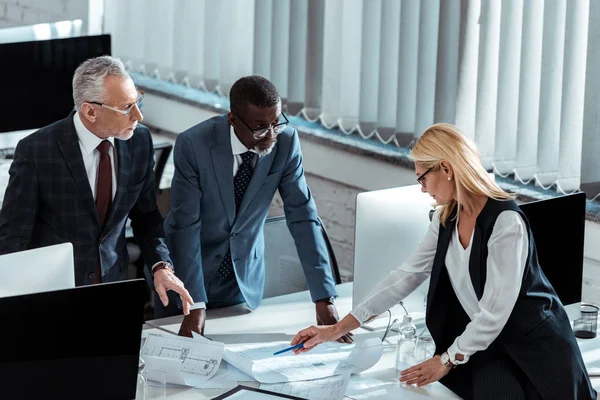 Overhead view of businesswoman in glasses holding pen near blueprints and multicultural businessmen — Stock Photo