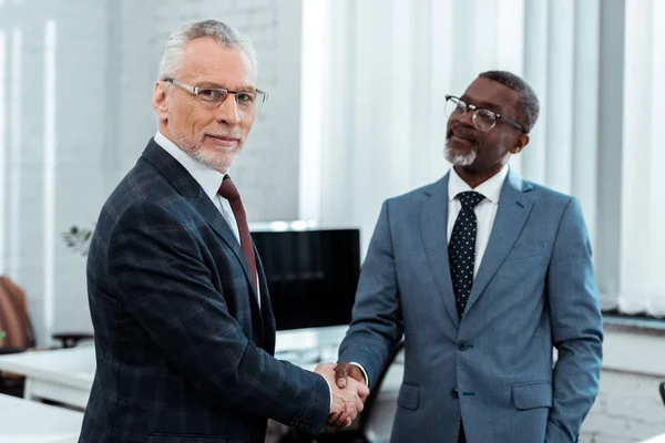 Enfoque selectivo de empresario feliz en gafas estrechando la mano con socio afroamericano - foto de stock