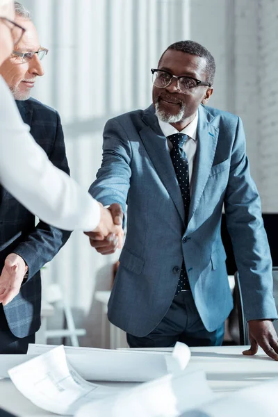 Selektiver Fokus des Geschäftsmannes mit Brille, der einen afrikanisch-amerikanischen Mann beim Händeschütteln mit einer Frau im Amt beobachtet — Stockfoto