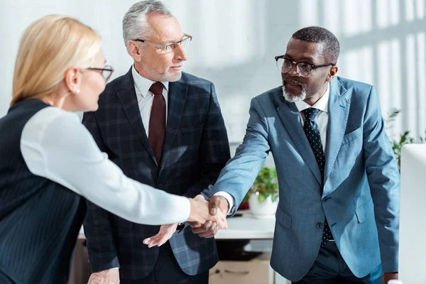 Enfoque selectivo de hombre de negocios en gafas mirando al hombre afroamericano guapo estrechando la mano con la mujer rubia en la oficina - foto de stock
