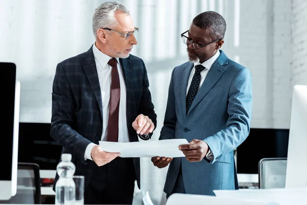 Handsome businessman in suit looking at african american partner while holding blueprints — Stock Photo