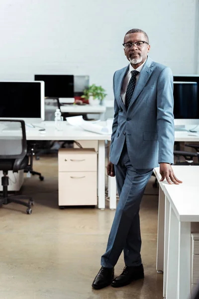 Красивий афроамериканський чоловік в окулярах стоїть в костюмі в сучасному офісі — стокове фото
