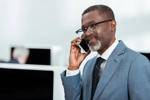 Guapo afroamericano hombre de negocios en traje hablando en teléfono inteligente en la oficina - foto de stock