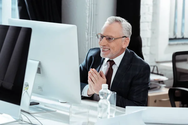Вибірковий фокус щасливого бізнесмена в окулярах жестикулює біля комп'ютерного монітора в сучасному офісі — стокове фото