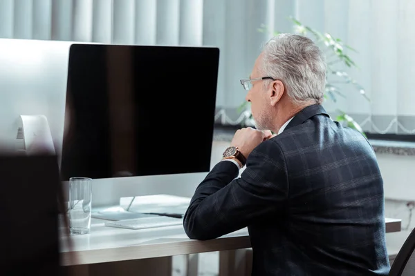 Selective focus of handsome businessman in glasses looking at computer monitor with blank screen in office — Stock Photo