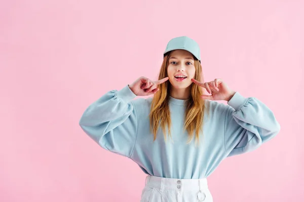 Bonita adolescente en gorra apuntando con los dedos a la sonrisa aislado en rosa - foto de stock