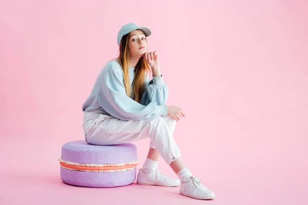 Pretty teenage girl in cap sitting on decorative macaroon on pink — Stock Photo