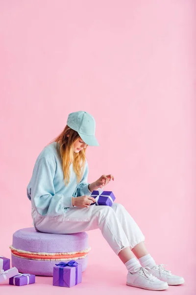 Pretty teenage girl in cap sitting on decorative macaroon and unpacking presents on pink — Stock Photo