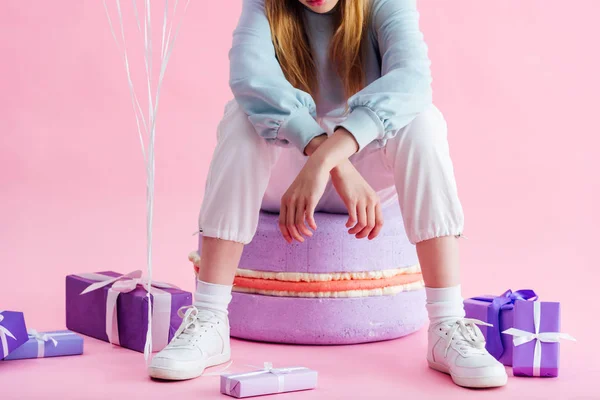 Vista recortada de la adolescente sentada en macarrón decorativo cerca de regalos en rosa - foto de stock
