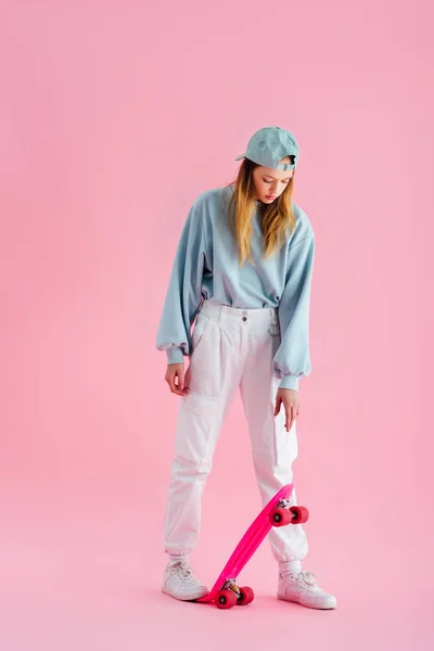 Pretty teenage girl in cap riding penny board on pink — Stock Photo
