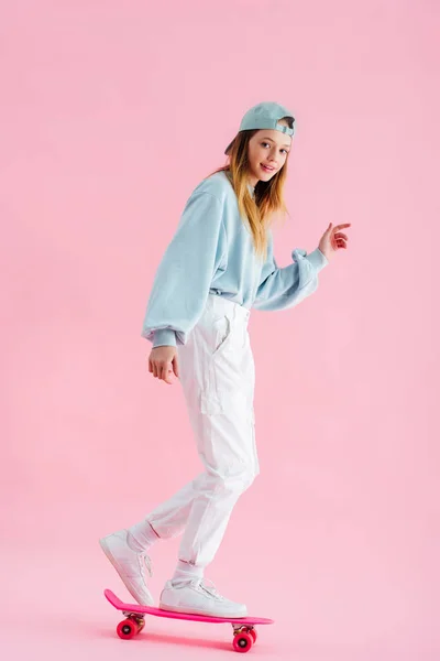 Happy pretty teenage girl in cap riding penny board on pink — Stock Photo