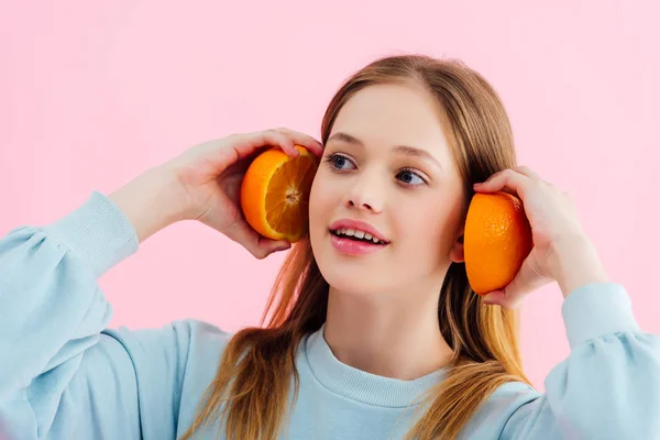 Sonriente bonita adolescente sosteniendo naranja mitades cerca de orejas aislado en rosa - foto de stock
