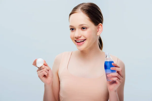 Happy smiling teenage girl holding cosmetic lotion and looking away isolated on grey — Stock Photo