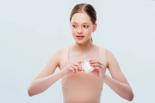 Adolescente sonriente aplicando crema cosmética en los dedos y mirando hacia otro lado aislado en gris - foto de stock