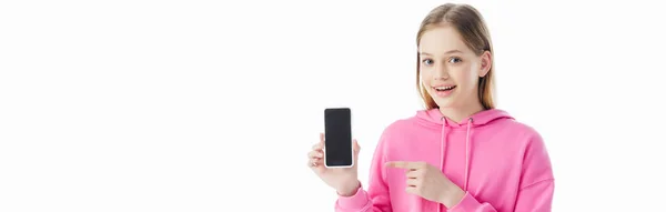 Panoramic shot of happy teenage girl in pink hoodie pointing with finger at smartphone isolated on white — Stock Photo