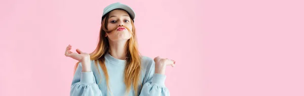 Colpo panoramico di bella adolescente in berretto con baffi di capelli sghignazzanti isolati su rosa — Foto stock