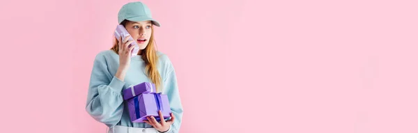 Панорамний знімок здивованої красивої дівчини-підлітка в шапці, що тримає подарунок, як телефон ізольований на рожевому — стокове фото