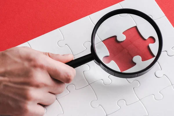 Cropped view of man holding magnifying glass near jigsaw shape and white connected puzzle pieces on red — Stock Photo