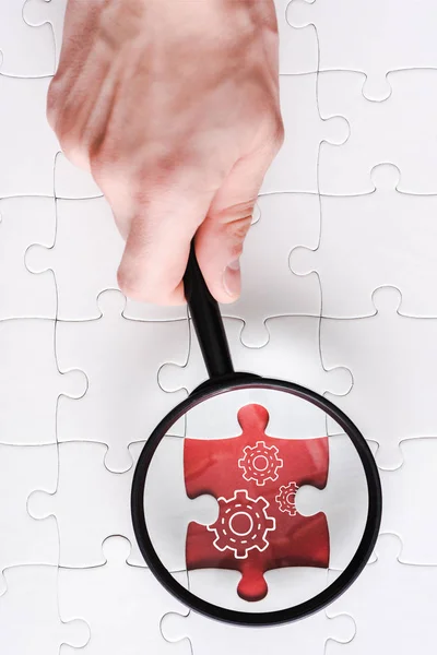 Top view of man holding magnifying glass near jigsaw with gears settings near white connected puzzle pieces — Stock Photo