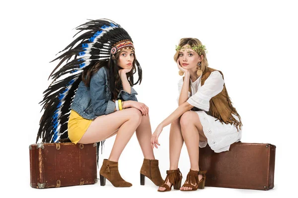 Due ragazze hippie bisessuali annoiate in copricapo indiano e corona seduta su valigie isolate su bianco — Foto stock