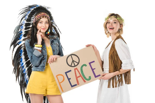 Deux filles hippies bisexuelles souriantes en coiffe indienne et couronne tenant une pancarte avec inscription isolée sur blanc — Stock Photo